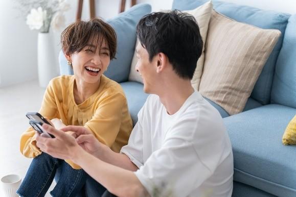 ソファーに座りスマホを見ながら笑顔で話す男女の画像